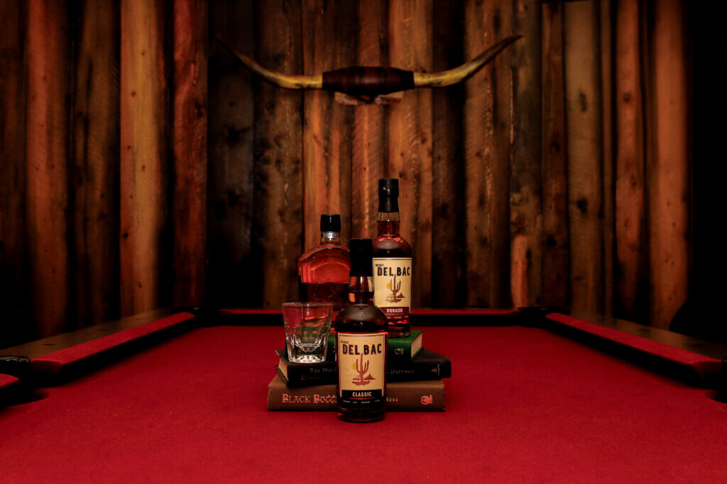 Whiskey Del Bac bottles and a tasting glass displayed on a red pool table beneath mounted longhorn horns at White Stallion Ranch.