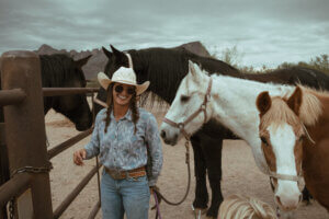 cowgirl with brunette hair braided in two in a cowboy hat smiling and leadind a white horse and a sorrel horse in front of a black horse behind a fence